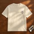 An off-white short-sleeve crewneck t-shirt with a small graphic on the left chest, laid flat on a wooden surface with dappled sunlight and a vintage cassette tape nearby.